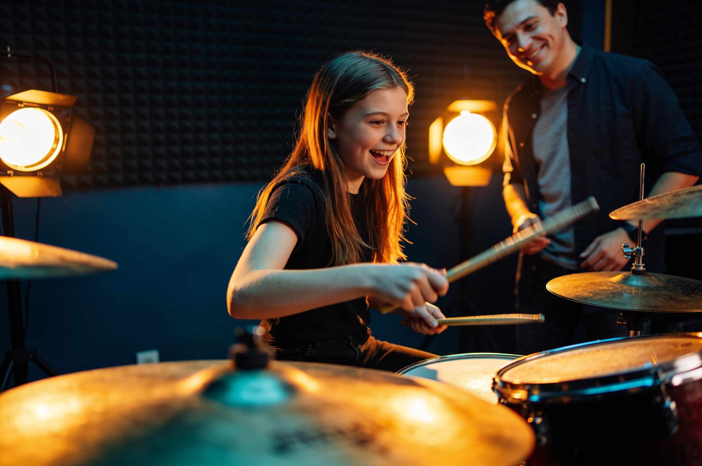 A teenage girl playing a drum kit with energy in an amber-lit practice room, her teacher nodding behind her.