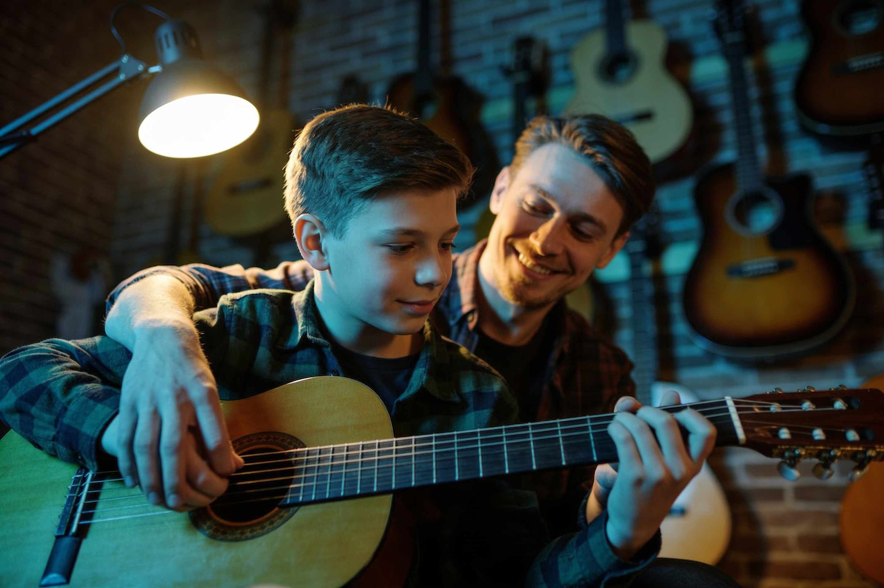 A teenage boy learning acoustic guitar with a smiling teacher beside him, guitars hanging on a brick wall behind.