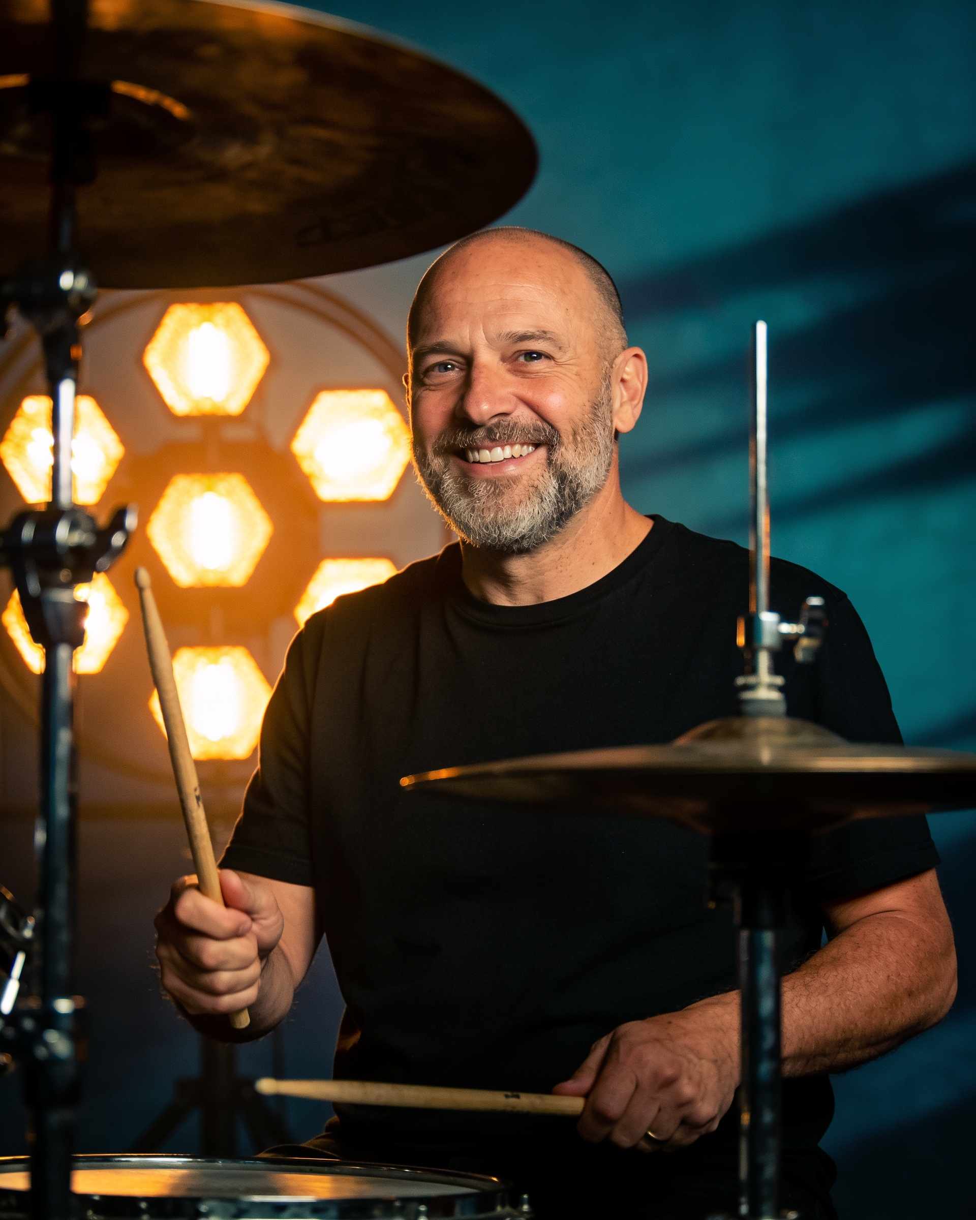 Portrait of a cheerful bald man with a salt-and-pepper beard holding drumsticks in front of a drum kit.