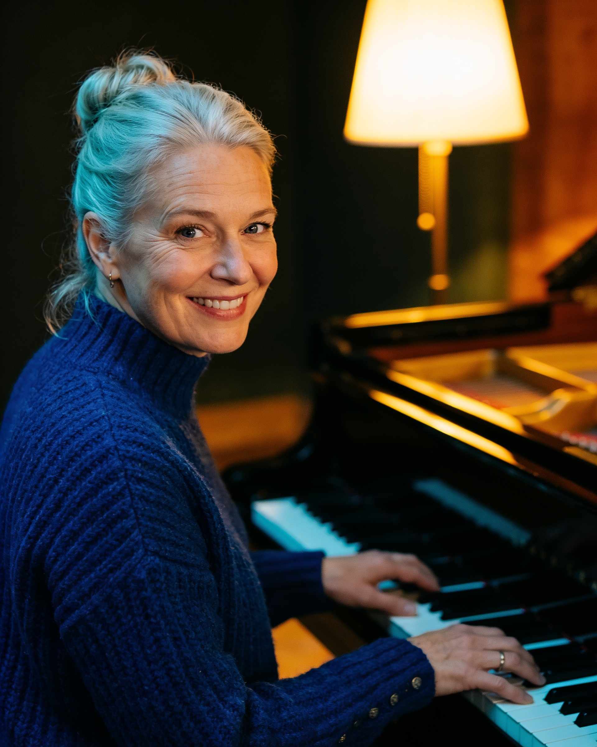 Portrait of a smiling woman with silver-blonde hair in a deep blue sweater at a grand piano.