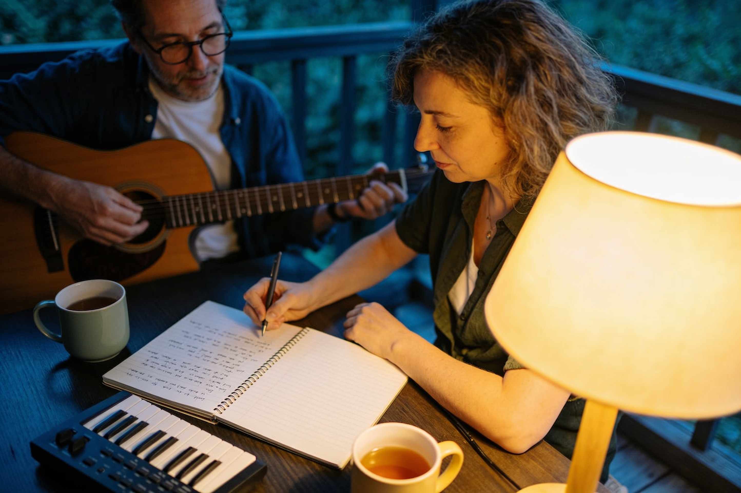 An adult student and a teacher writing a song at a table with a lyric notebook, a guitar and a small keyboard under a lamp.