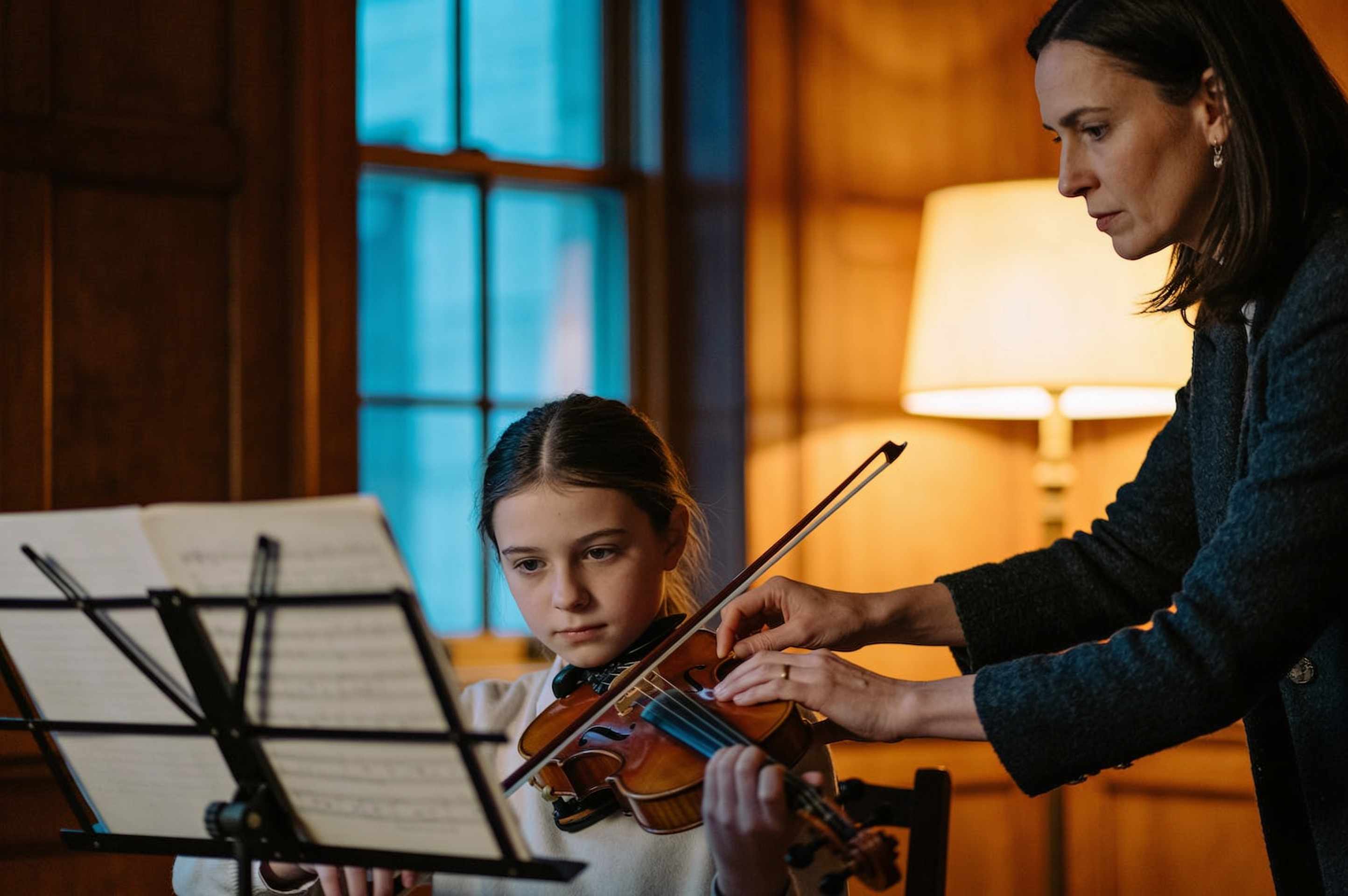 A girl playing violin at a music stand while her teacher guides her bow arm in a warm wood-panelled room.