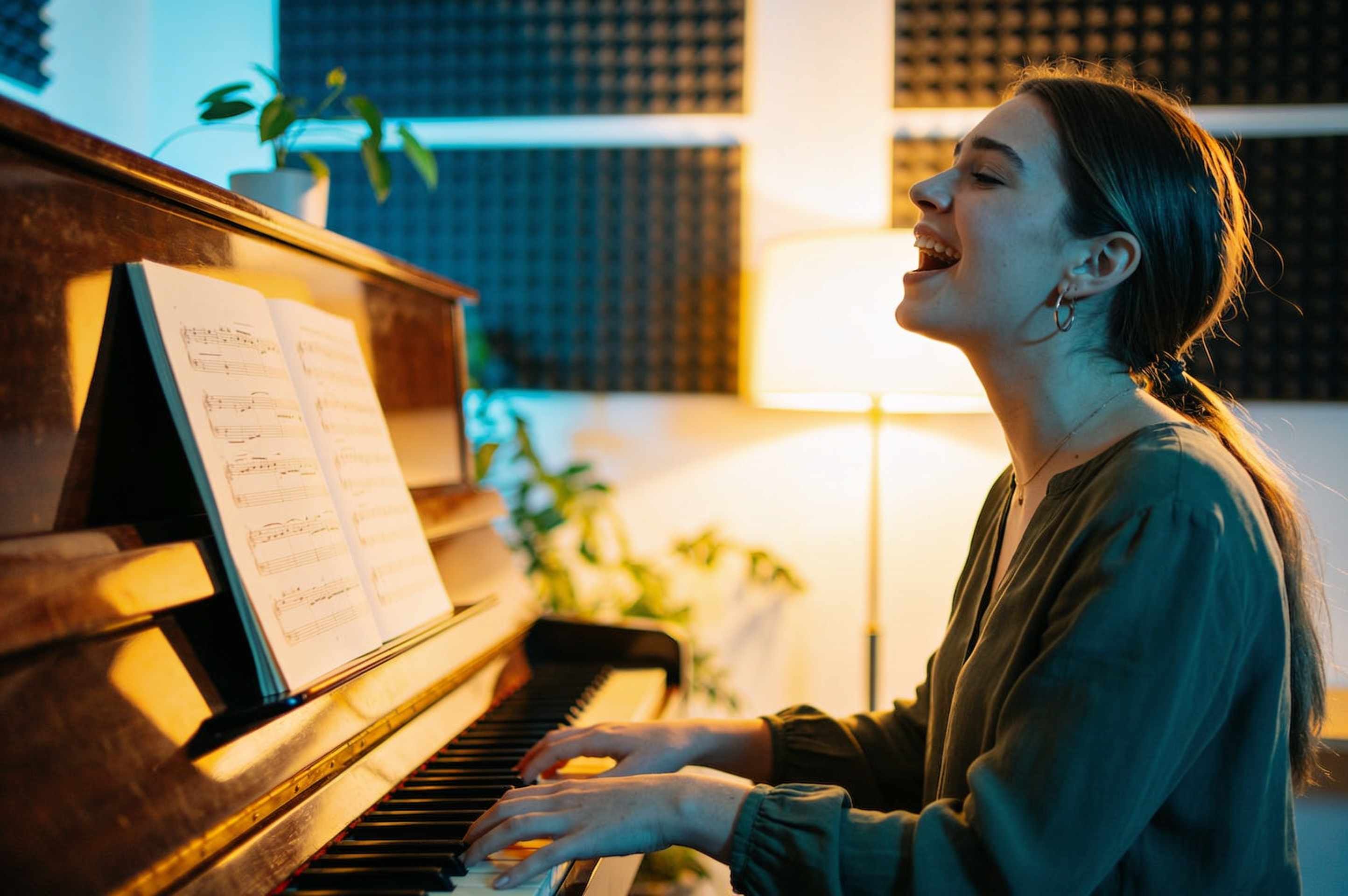 A young woman singing joyfully beside an upright piano in a bright studio with plants.