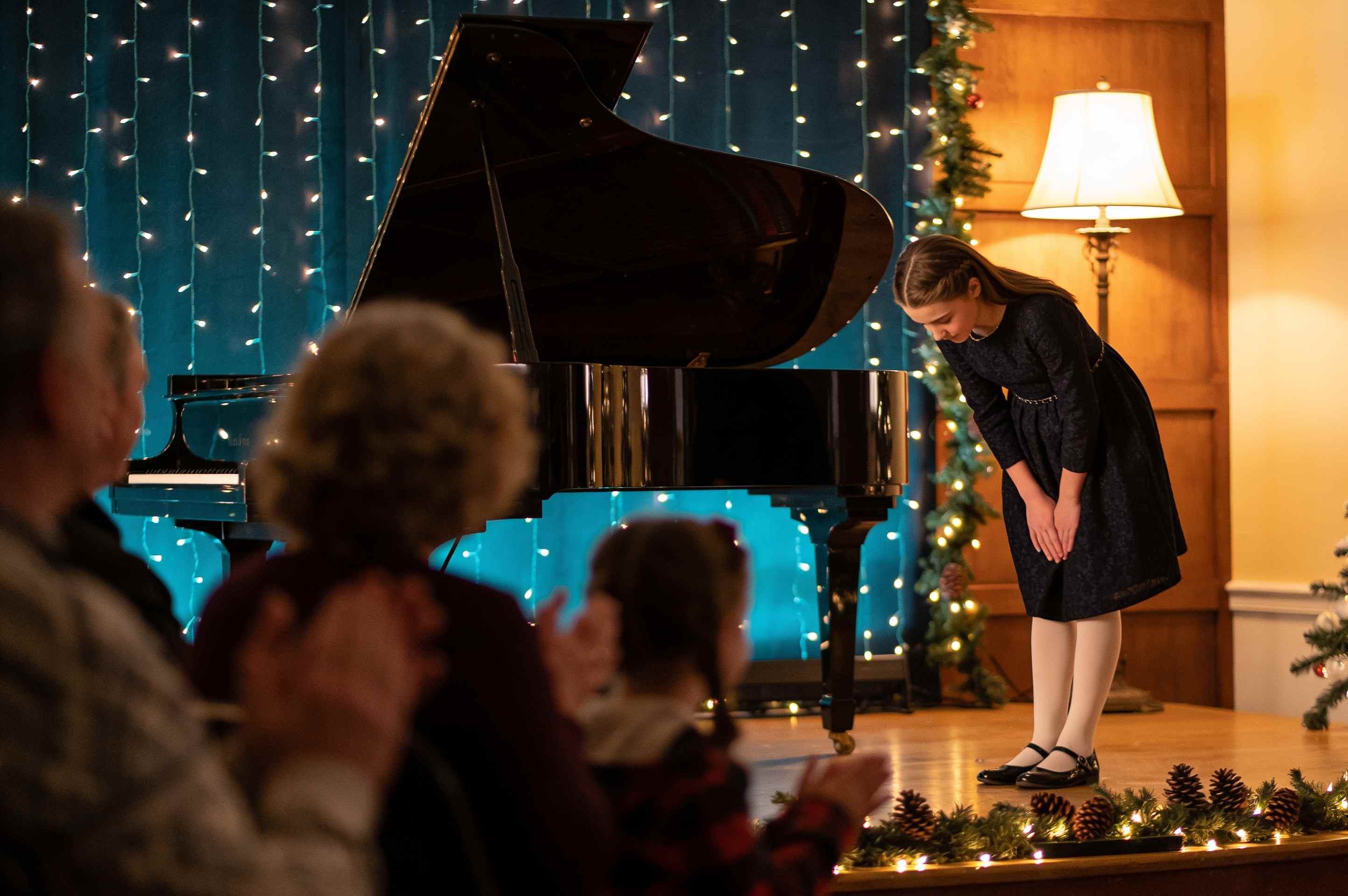 A young pianist bowing on a small stage beside a grand piano at a winter recital with string lights and pine garlands.